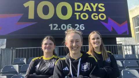 Three Gloucestershire county cricket club players pose for a photo in front of a large screen which has '100 days to go' in large yellow text on it to mark the countdown to the women's T20 world cup starting. All three players and smiling at the camera and wearing black Gloucestershire kit with yellow stripes across the shoulders.