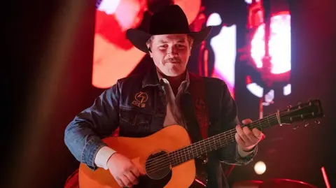 Getty Images Zach Top wearing a black stetson hat and blue denim jacket plays an acoustic guitar. He is standing on a stage with a red background.