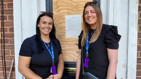 Two female council officers, wearing lanyards, stand outside a front door which has a typed sheet of A4 paper stuck to it.