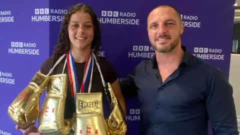 A young girl with dark brown curly hair is smiling at the camera and holding up both her fists. She is wearing a black top and around her neck are three gold boxing gloves and a gold medal with blue, white and red stripes on the ribbon. In the background is a purple wall which repeatedly says 'BBC Radio Humberside'. Next to her is a man with a shaved head who is smiling and wearing a navy long-sleeved shirt.