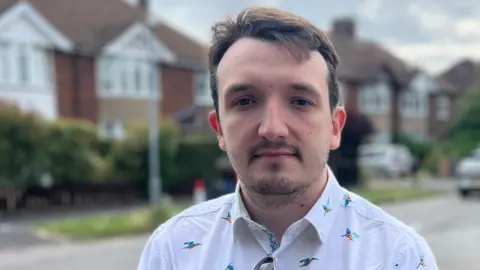 Tom Jackson/BBC A head and shoulders picture of a man with short dark hair and dark stubble. He is wearing a shirt with small kingfishers on it. He is standing in a residential street.