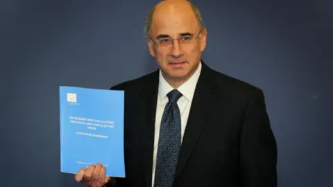 PA Media Lord Leveson is pictured in 2012 holding a copy of his report which scrutinised how the media in the UK operates. He is wearing a dark suit, white shirt and a dark blue tie as he looks ahead. 