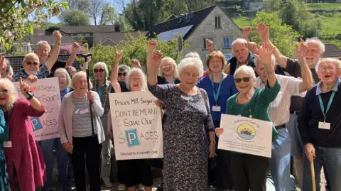 A large group of elderly people are all posing for a photo. Two are holding up signs that say "Price's Mill Surgery, don't be mean, save our spaces". They are all cheering in the photo which is taken outside in a rural area with the Price's Mill Surgery in the background. 
