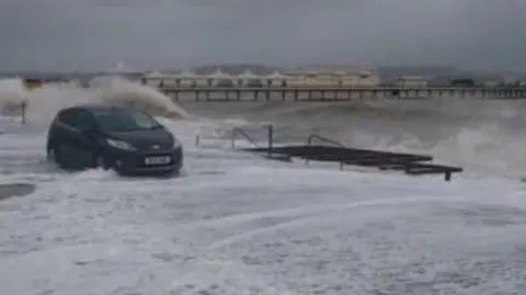 Torbay Council A black car is pounded by the waves on Paignton seafront during Storm Emma in March 2018. You can't see the beach for all the foam. The pier is in the distance set against a stormy grey sky.