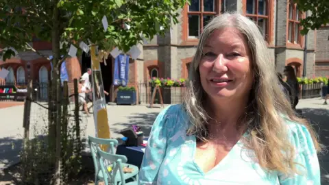 Sinead Gibson is wearing a light blue dress, has long grey hair and is stood outside Reading Town Hall.
