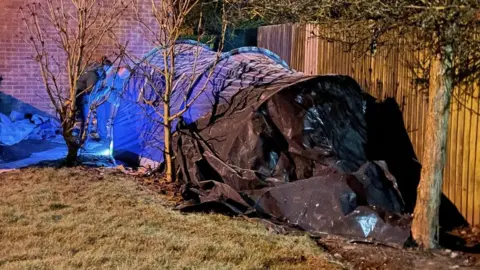 A large blue tent is pitched next to a garden fence