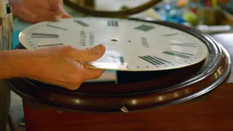 The Repair Shop Round brown wooden clock laid down on a table - its face is being lifted out by a man - his hands are holding the face on each side.