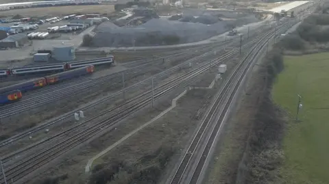 Mark Dodd/BBC An aerial view of Ely junction showing different trackes converging on a double set of rails in the distance. Three stationary trains can be seen on sidings on the far right of the picture. There are piles of aggregate to the left of the railway lines.