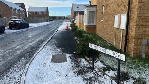 A street sign reading Mitford Grove. There are rows of semi-detached brown brick houses on both sides of the road. An orange cone is put on the pavement. There is a bit of snow and a parked car on the left side of the street. 