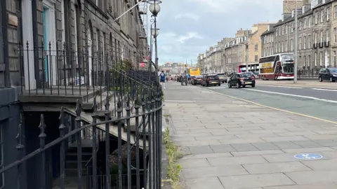 A street with buses and cars on the road, a wide stone slab pavement, and railings and an area in shadown below steps to the left.