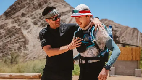 A man puts his arm around a runner who is carrying water bottles and is wearing a sun hat with sunglasses. A mountain and desert scrub can be seen behind him.