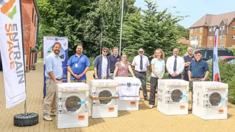 Entrain Space People standing in a residential area for a picture with five washer-dryers in front of them and charity banners