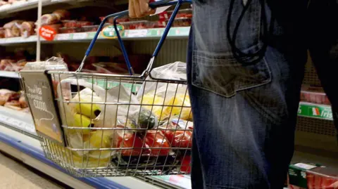 A basket of shopping being carried by a man in a supermarket. It has fruit and vegetables in it. You can only see part of the man's hand and he is wearing jeans. He is stood in front of a fresh meat counter