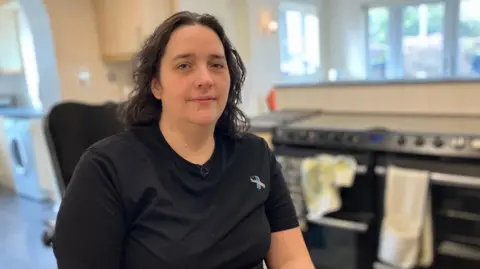 Susannah Thompson has long dark hair and a black t-shirt with a ribbon logo. She is sat in a kitchen.
