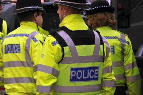 A rear view of three West Midlands Police officers - two male and one female - all wearing bright yellow high-vis police jackets and wearing helmets. 