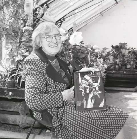 Hull University Archives A black and white image of Ms Crackles holding her book, she is seated in a greenhouse and is wearing a skirt and jacket in spotted fabric.