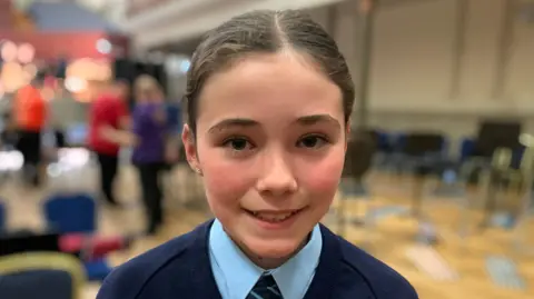 A girl with long brown hair pulled away from her face is looking at the camera smiling. She is wearing a navy v necked jumper over a pale blue shirt with a navy and pale blue tie.