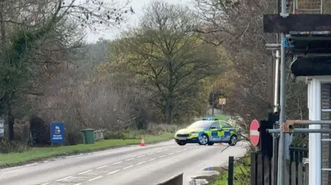A road surrounded by trees with a police car parked on the road cordoning it off.