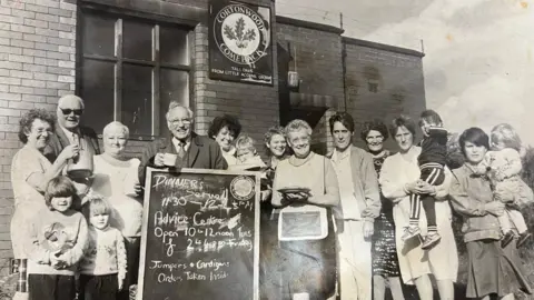 Cortonwood Comeback A group of people, mostly women, stand in a line in front of a brick building. They are all smiling and some are holding children or cups of tea or food. The photo is in black and white.