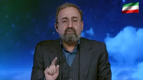 A TV presenter speaking to camera with his finger raised in front of a blue cloudy backdrop and the Iranian flag in the top right corner