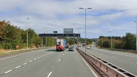 Google maps screengrab of the A55 at Dwygyfylchi, Conwy.  Cars can be seen travelling away from the camera. Blue skies can be seen as green hedges line the road. 