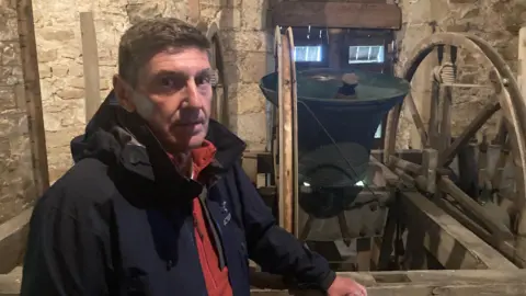 A man stands inside what appears to be an old stone bell tower. The walls around him are built from large, weathered blocks of stone, giving the space a distinctly medieval or early‑historic feel.