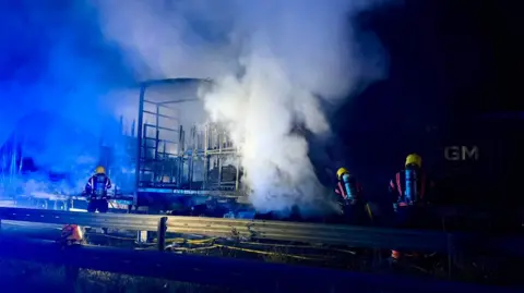 Peterborough Volunteer Fire Brigade Smoke rises from a burned out lorry, with fire crews tackling the blaze in the dark.