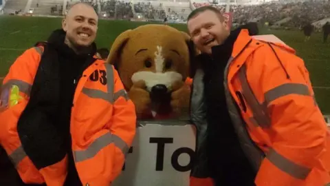 Ben Moss Ben Moss and Tom Doolan are smiling while wearing hi-vis orange jackets, with a football mascot that looks like a dog standing between them, at a football ground