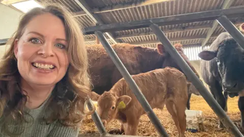 Brook Meadow Campsite Image shows Claire smiling and kneeling down in front of the pen with the cows in. The calf is at the front and the mother cow is black and on the right next to the small calf. The other calf is a bit bigger and is standing behind the smaller newborn one and is also brown. Claire has long blonde/brown hair and blue eyes. She's wearing a light blue ribbed jumper. 