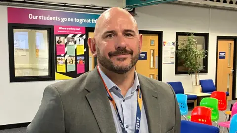 A bald man with a dark chin beard wearing a blue shirt and grey jacket standing in the college foyer.