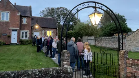 People queue outside a village hall for a public meeting. An old wrought iron arch rises over the gates, and a green lawn flanks the path up to the village hall