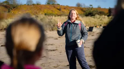 Dru Dodd Bridie Melkerts is talking to a group of children on a beach with the image taken from behind the heads of two of the children. She is wearing jeans and a blue top and has long blonde hair. 