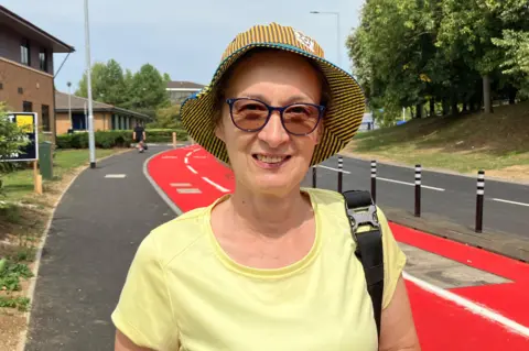 Shariqua Ahmed/BBC Sue wearing a yellow T-shirt and a sun hat - smiling for the camera. The red cycle path stretches behind her.