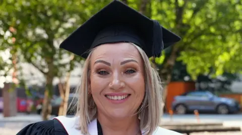 Rachel, a woman with straight shoulder-length blonde hair wearing a graduation cap. She has a nose stud, and wears light pink lipstick. 