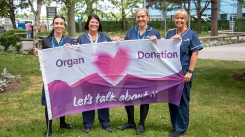 Four organ donation nurses in blue uniforms hold the flag outside of the Hull Royal Infirmary. The flag is purple and white and has a heart in the middle with the words Organ Donation Let's Talk About It.