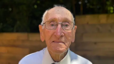 George King/BBC A head and shoulders image of an elderly man wearing a white shirt and glasses. He is looking directly into the camera.
