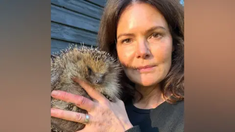 Contributed A woman with short brown hair holds a large hedgehog in her hands as she looks and smiles slightly at the camera. There is a silver ring on one of her fingers. 