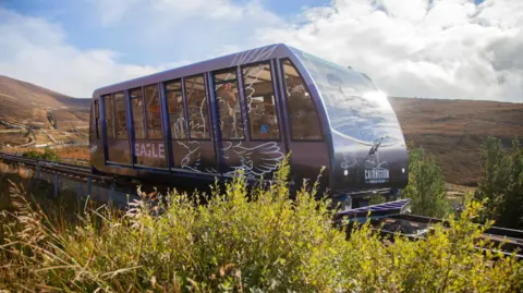 One of the funicular's vehicles travels along the rails on a sunny day. The carriage is a dark purple, similar to the colour of flowering heather. The vehicle, which is passing an area of green vegetation, has the word "eagle" and a stylised drawing of an eagle on the side.