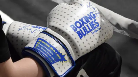 Vicky Davies The image shows a white and blue boxing glove with grey stars, Boxing Bullies by Jake Paul is written on the front of the glove in blue writing.