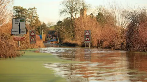 Weather Watchers/Cloud9Weather A view of a flooded road with brown trees around and Glastonbury Tor in the background.