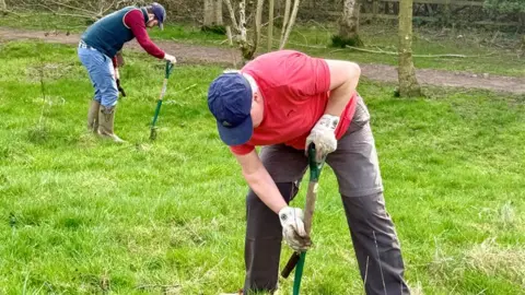 Two people wearing outdoor clothing use spades to dig into the grass in a wooded area, with trees and a path visible in the background.