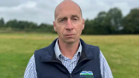 A bald man wearing a blue NFU Scotland gilet over a blue and white checked shirt. He is staring into the camera and standing in a field with a row of trees behind him.