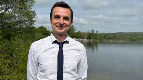 Howard Perry - a man with short dark hair, a black tie and white shirt, stands and smiles. A tree is right behind him, as is a large reservoir. With the far-side shore visible in the distance.
