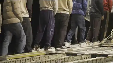 The US Seema Bal still walking on a ramp on a military aircraft, showing migrants in a casual cloth from a video made by a video made by the US border force and holding a warm coat together with their feet. The image is cropped to hide their face and highlight chains around their ankles.