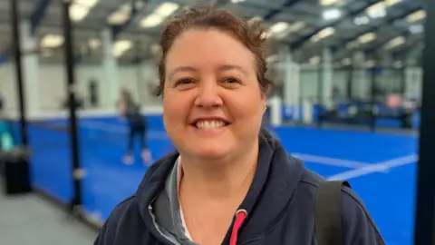 Sam Read/BBC A woman with brown hair tied back stands against the backdrop of a padel court where a match is being played.