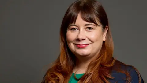 Parliament Anneliese Midgley has long brown hair and a split fringe. She wears a green dress and a blue jacket. She smiles at the camera