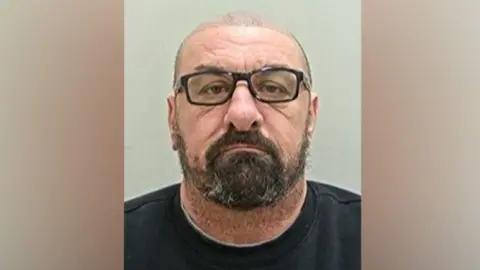 Christopher O'Neill's head and shoulders mugshot. He has shaved hair, black-rimmed glasses and a black beard. He is wearing a black t-shirt and is in front of a grey background