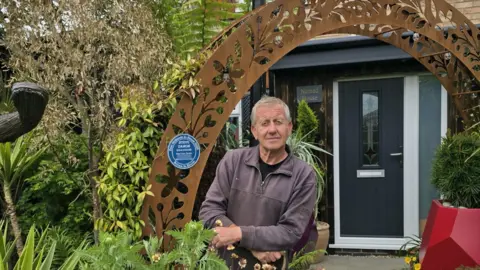 Steve Damon is pictured in his front garden. He is standing under an ornate rusted metal archway on which there is a blue plaque which reads 'Steve Damon, gold award, best front garden, East Midlands in Bloom 2025'. The black front door of Steve's house can be seen just beyond the arch. The garden is filled with tropical plants and sculptures.