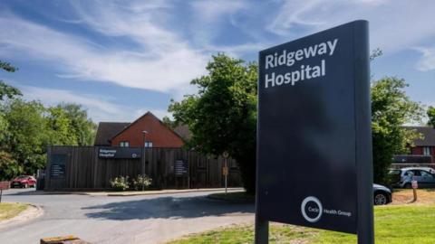 Ridgeway Hospital sign, set on grass, with hospital buildings behind it