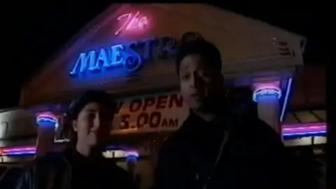 Moss Sheikh/Mick Chandsoor/Legacy Bhangra Daytimers Two young South Asian people standing in front of a building with a neon pink and blue sign saying 'The Maestro', and 'open until 3am'